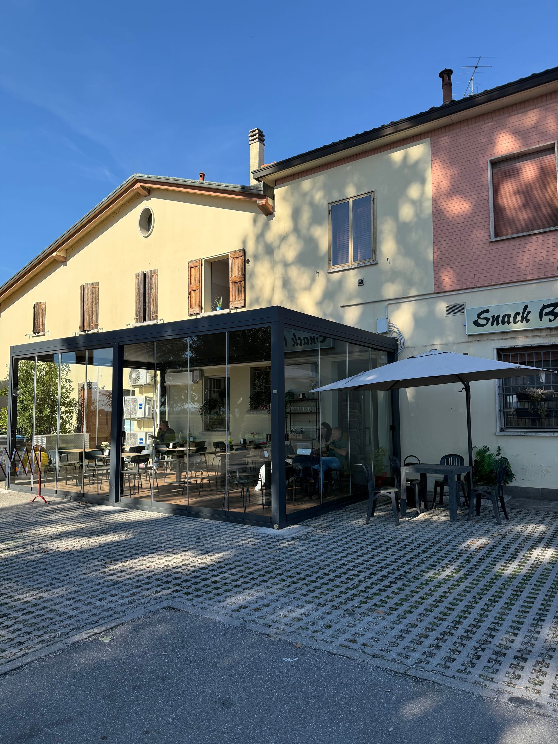 Bar con vetrate e posti a sedere all'aperto su un'area pavimentata, sotto un cielo azzurro.