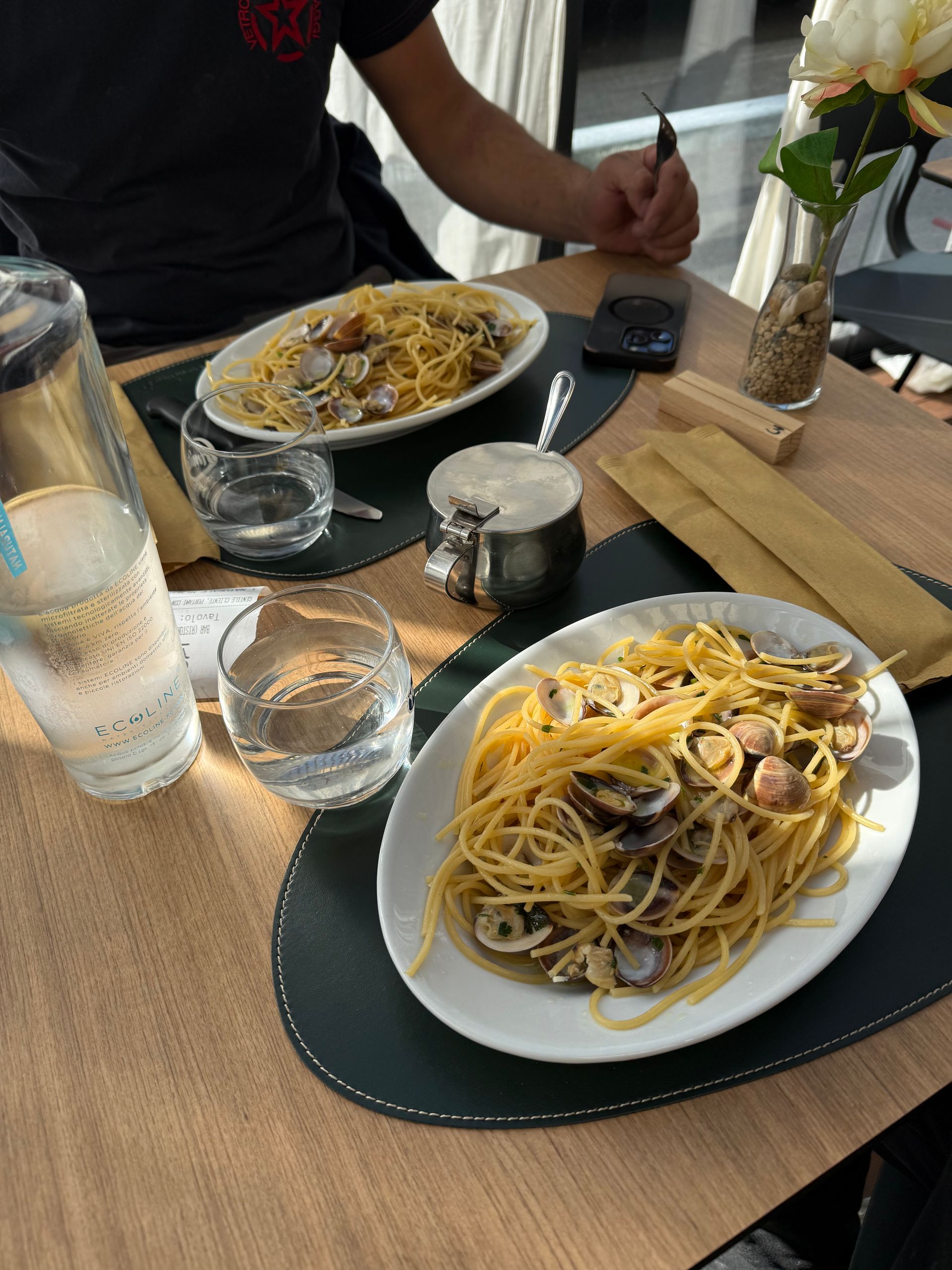 Due piatti di pasta con le vongole su un tavolo, bicchieri d'acqua, una persona seduta.