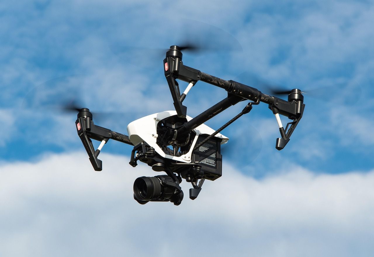 Drone in flight, white and black design, camera attached, against a cloudy blue sky.