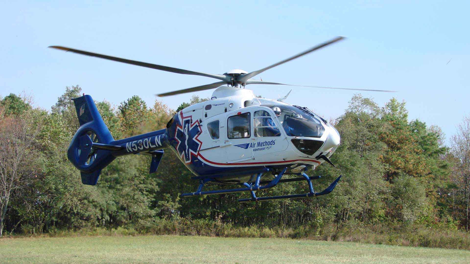 Medical helicopter with blue and red markings taking off over a green field and trees.