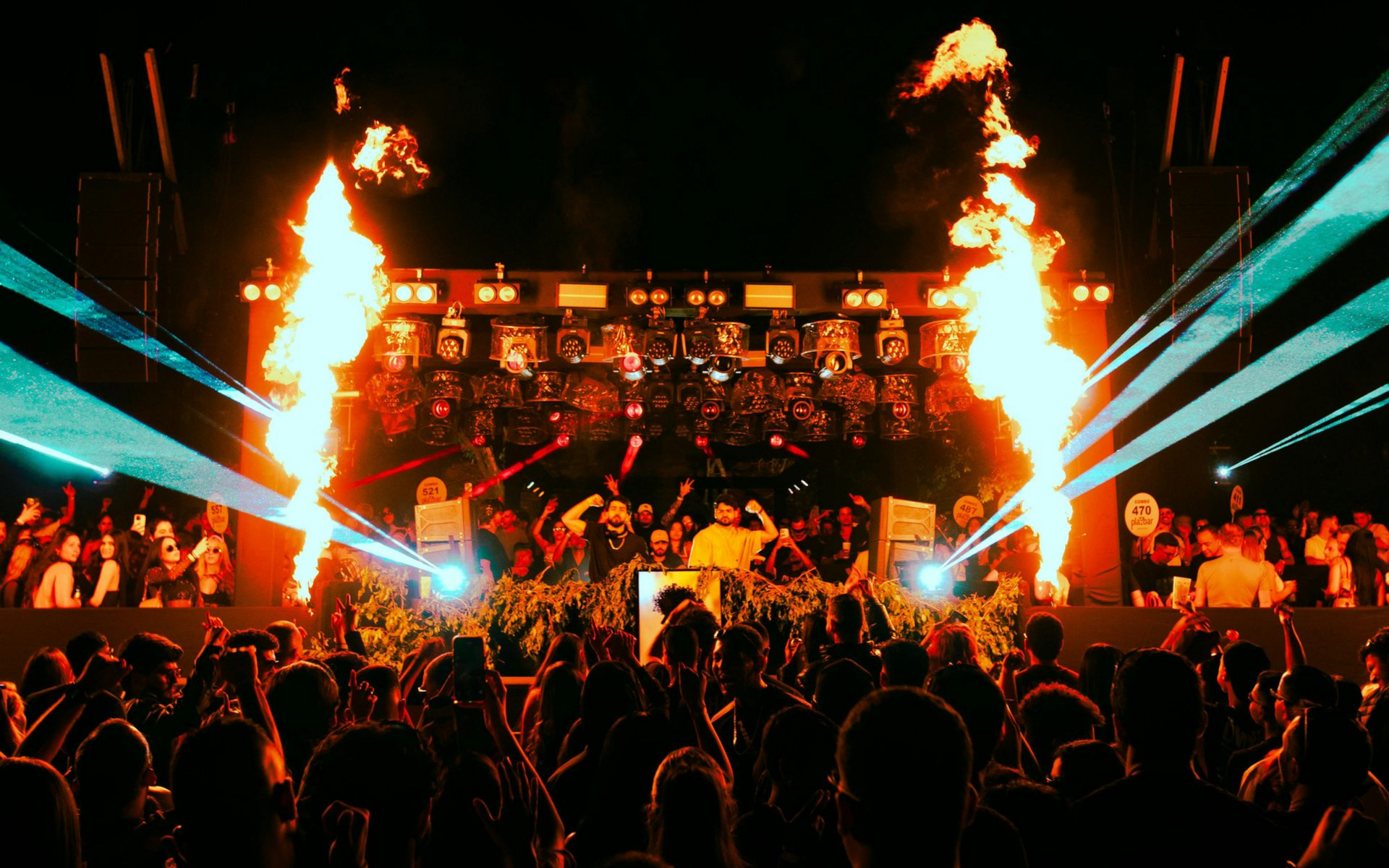 Concert with DJ on stage, pyrotechnics, and a crowd. Flames erupt, teal lights shine, and people dance. Concert with DJ on stage, pyrotechnics, and a crowd. Flames erupt, teal lights shine, and people dance.