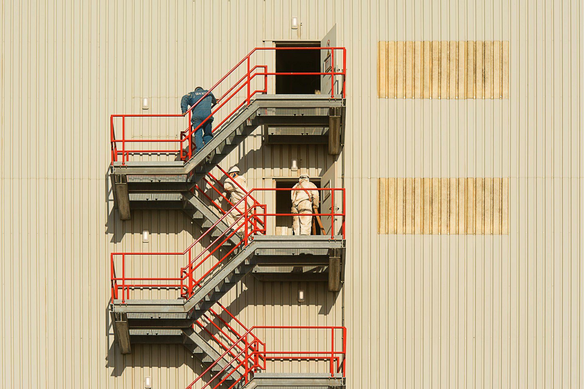 Workers in safety gear on a metal exterior staircase attached to a large, light-colored industrial wall.