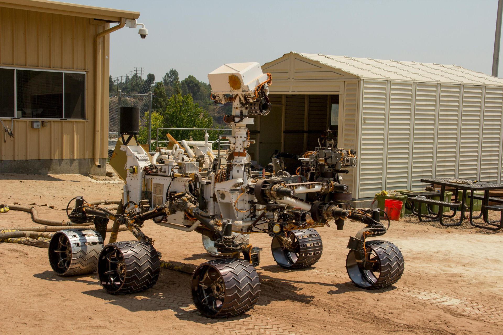 Mars rover on a dirt lot, near buildings. The rover has multiple instruments and wheels; sunlight.