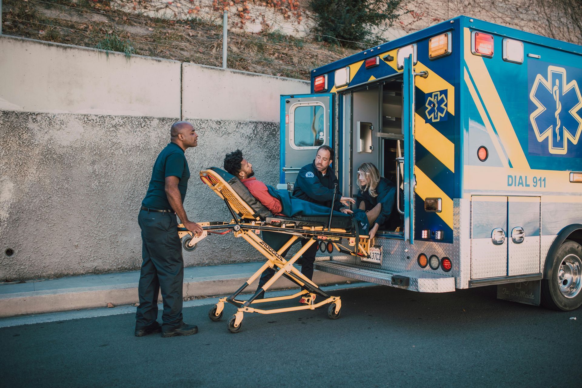 Two EMTs loading a patient on a stretcher into an ambulance; another EMT stands nearby.