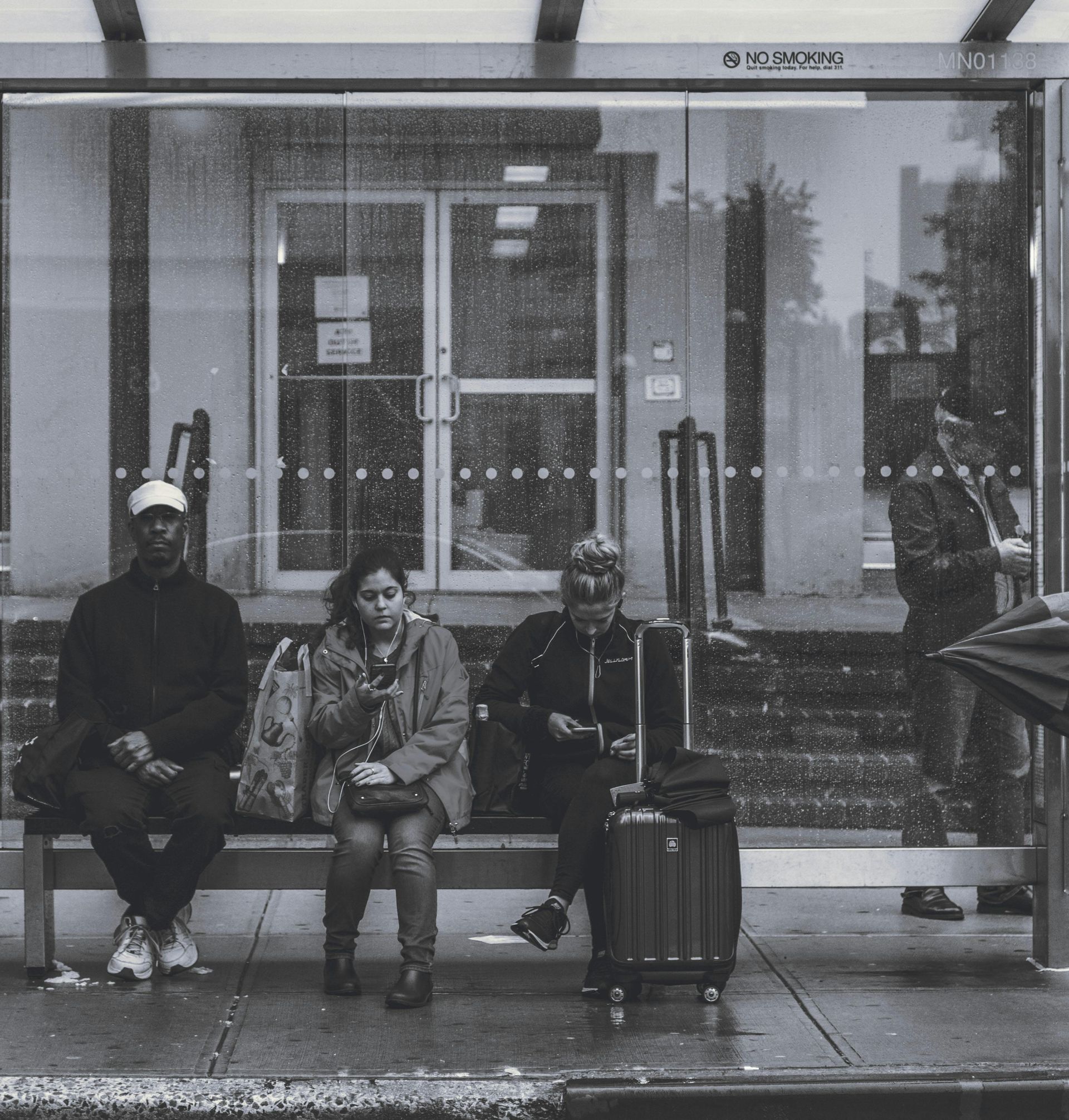 People waiting at a bus stop. Rain streaks the glass. A suitcase sits beside a person. One person looking at their for to see when the bus arrives with de Lijn app.