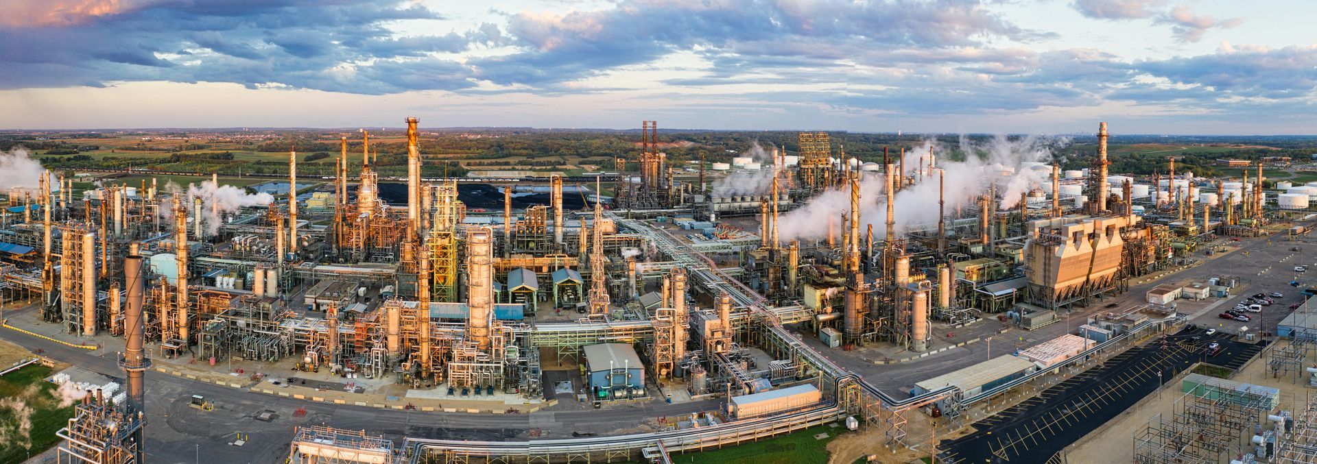A wide-angle aerial view of a large industrial oil refinery at sunset, featuring tall towers, pipes, and steam plumes.