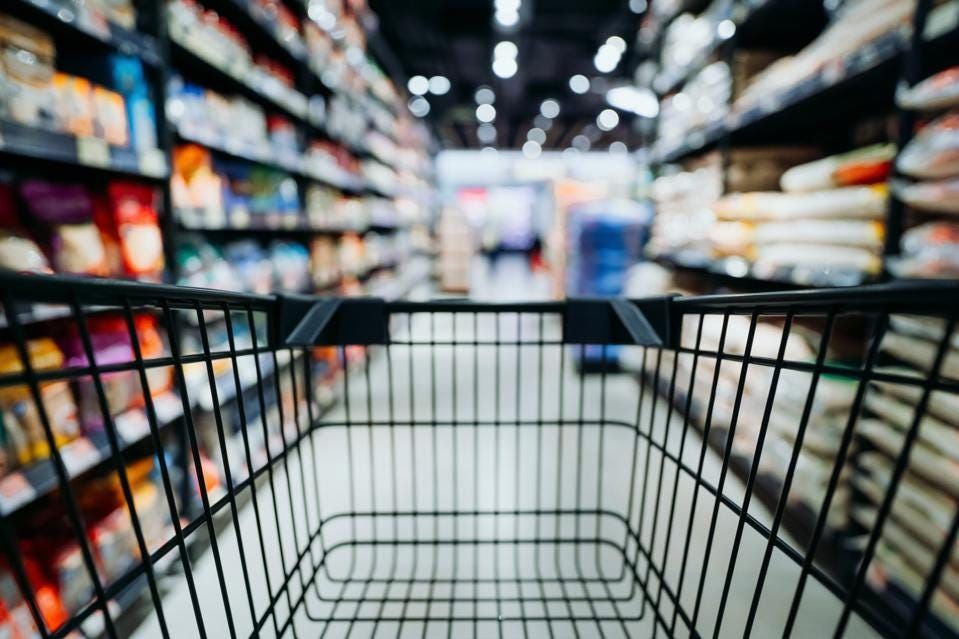 A first-person view looking through an empty black metal shopping cart in a brightly lit supermarket aisle.