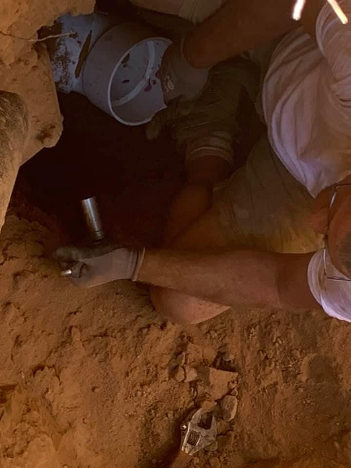 A man is kneeling down in the dirt holding a flashlight.