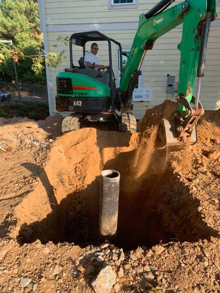 A green excavator is digging a hole in the dirt in front of a house.