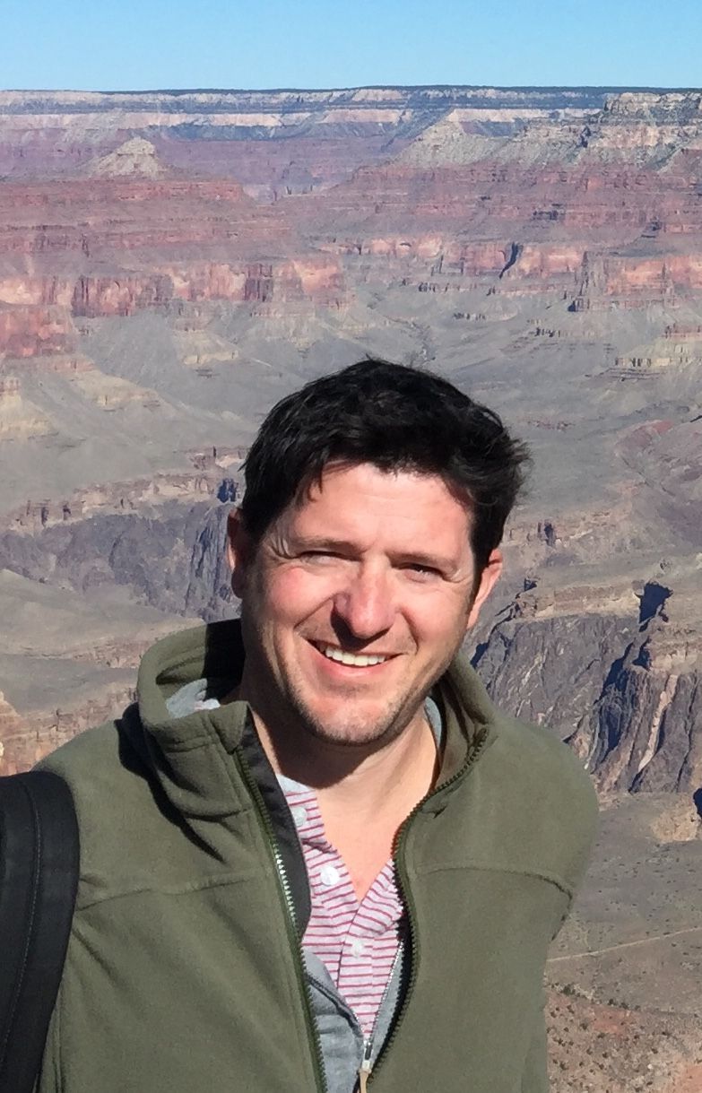 A man in a green jacket is standing in front of a canyon.
