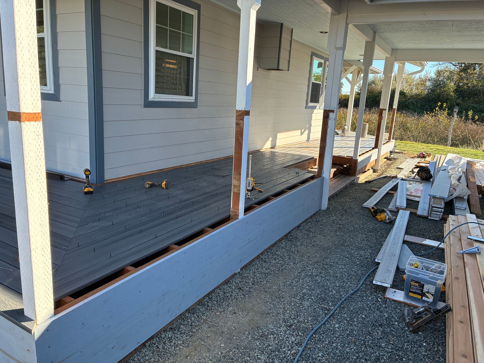 House porch under construction with white posts, blue skirting, gravel ground, and stacked materials along the side.