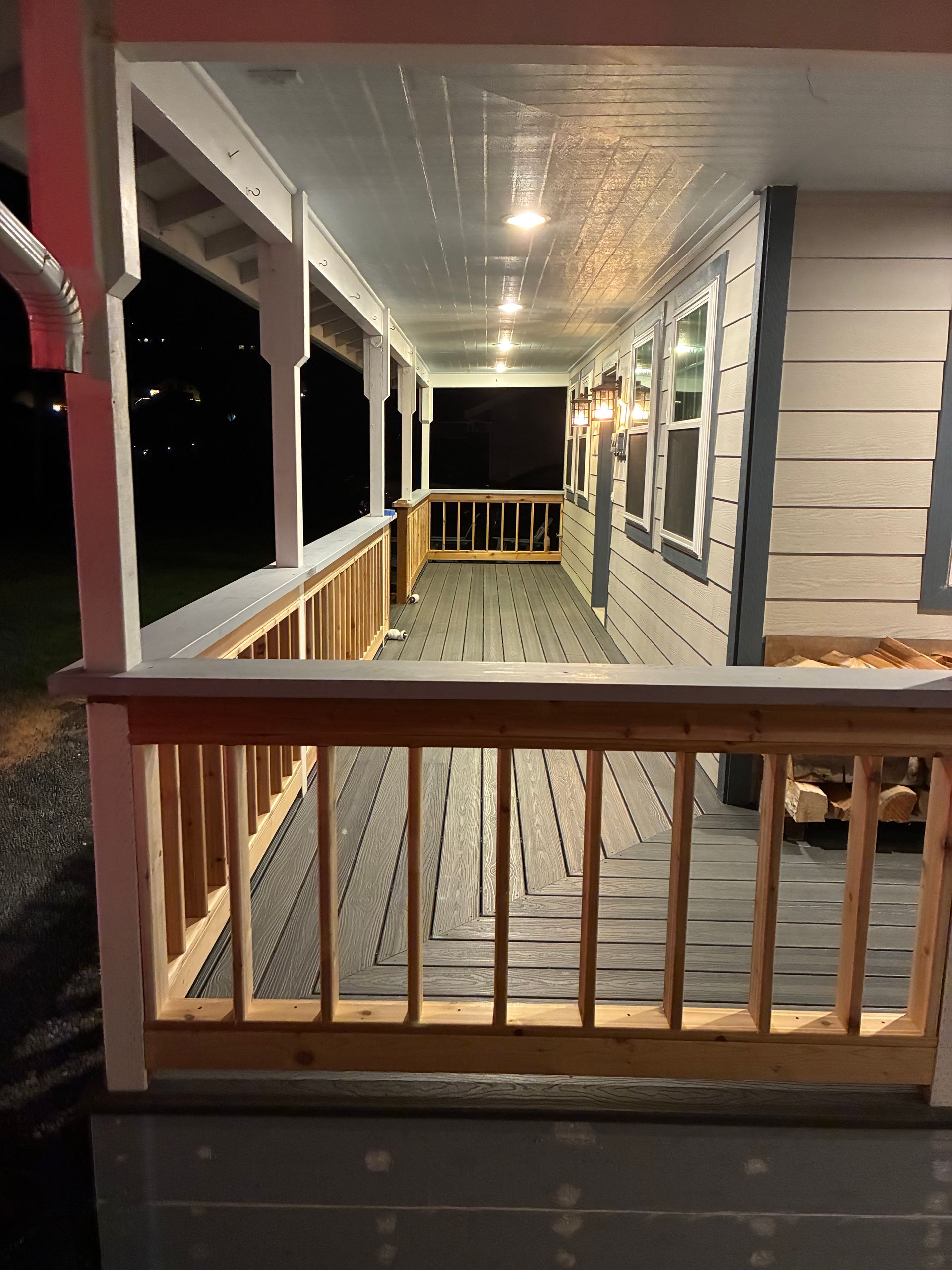 Covered front porch with wooden railing, gray floor, and windows along the house at night.