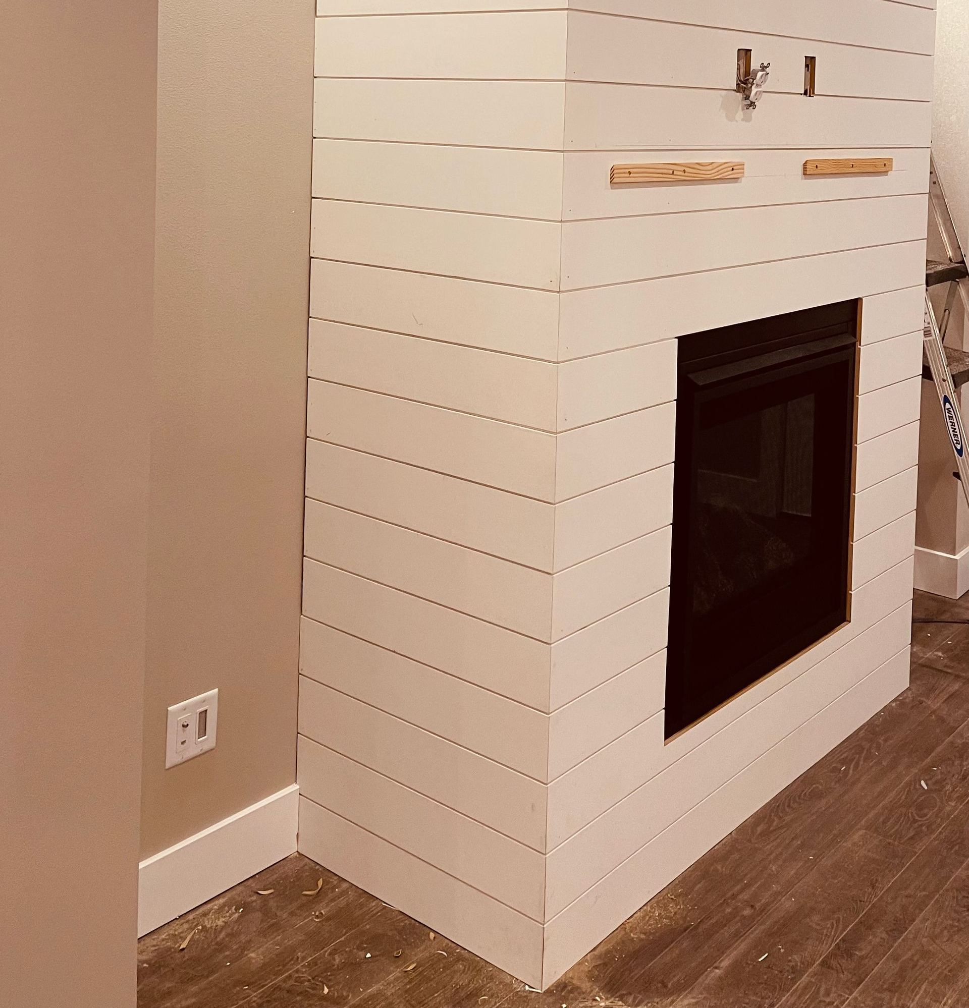 White tiled fireplace on a wood floor beside a beige wall and outlet.