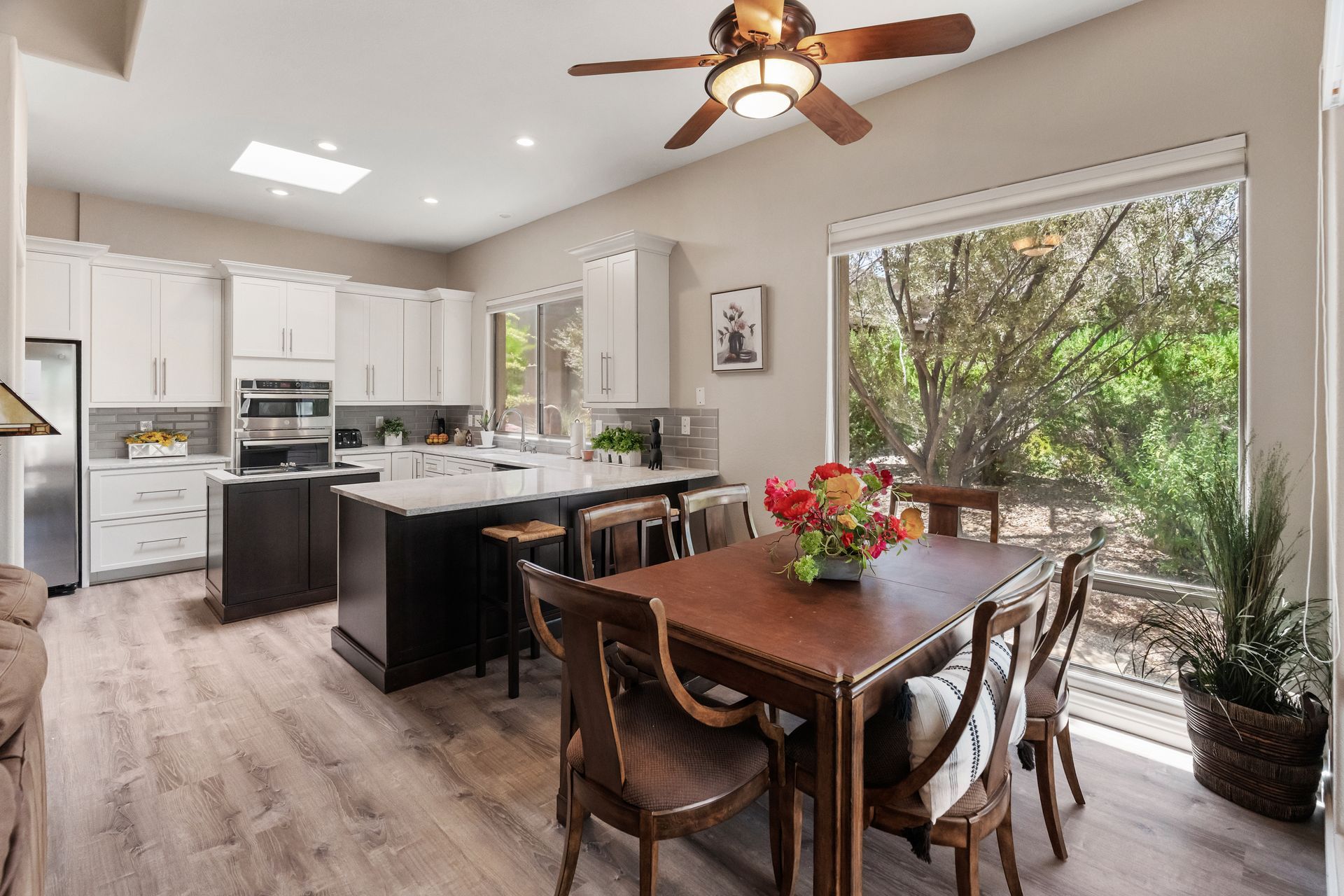 Kitchen with white cabinets, dark island, dining table, and large window overlooking trees.