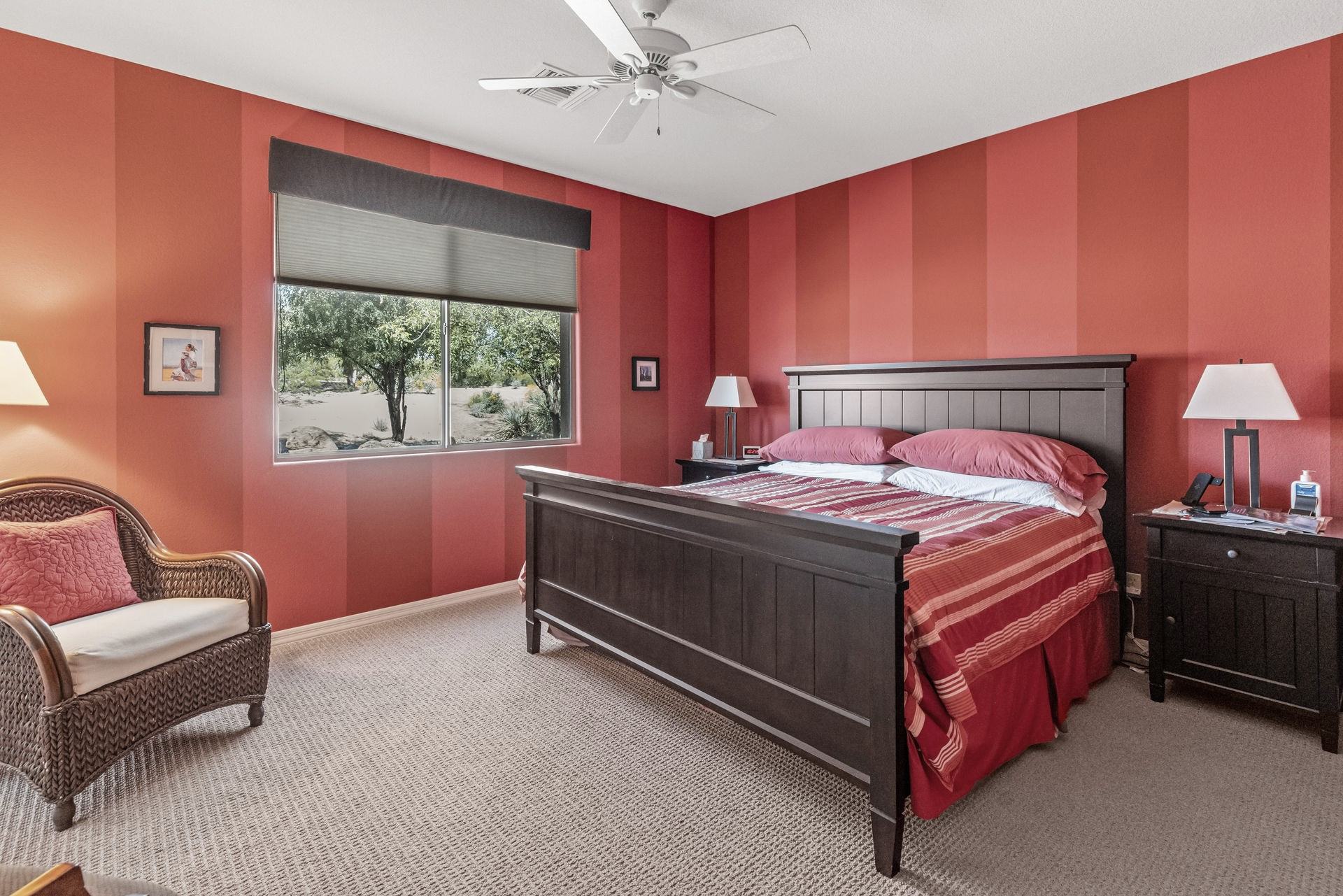 Bedroom with bed, wicker chair, window, ceiling fan, and art. Neutral colors, natural light.