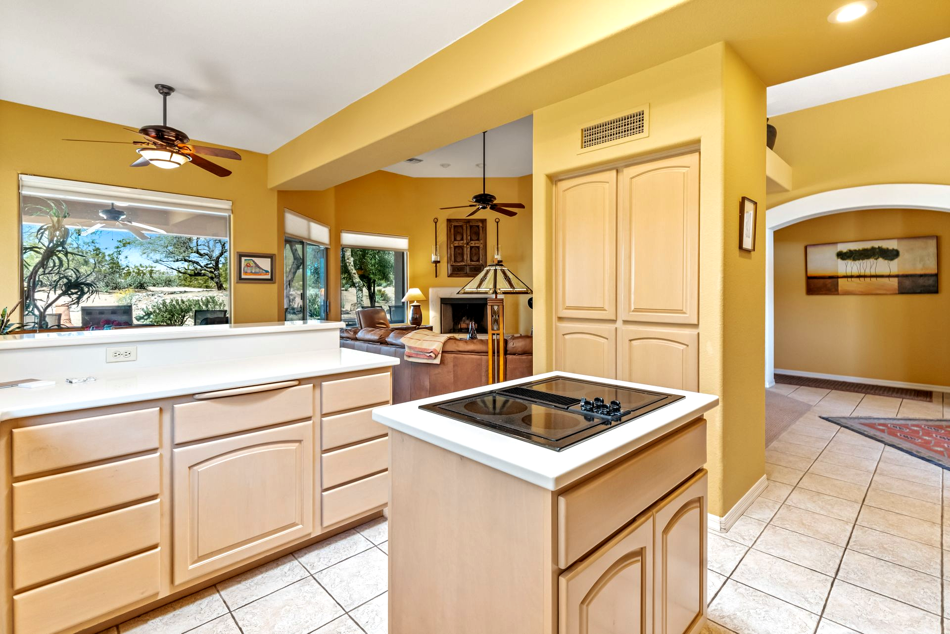 Kitchen with island, tan cabinets, and yellow walls. View to living room and outdoors.