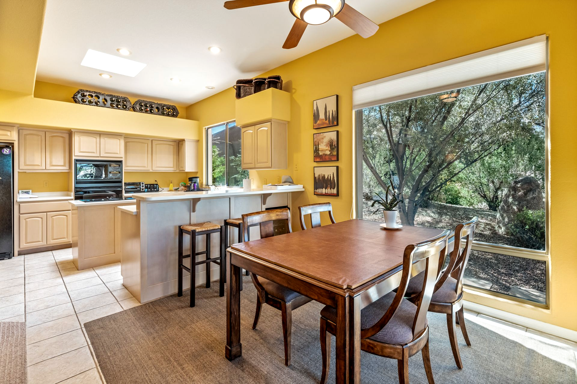 Yellow kitchen with wood table and chairs, tan cabinets, and a view of trees through a window.