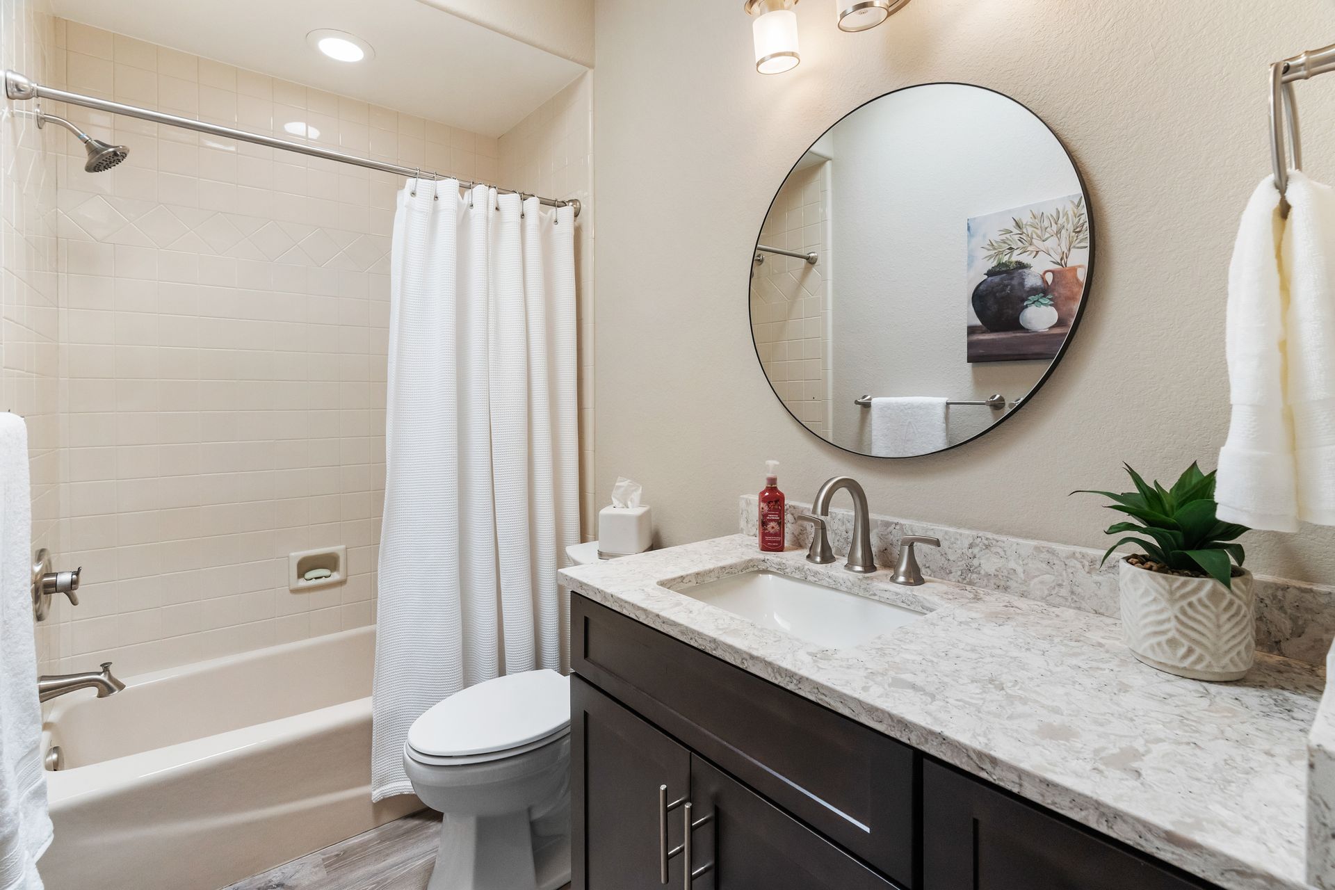 Bathroom with a dark brown vanity, round mirror, white sink, toilet, and a shower/tub combo.