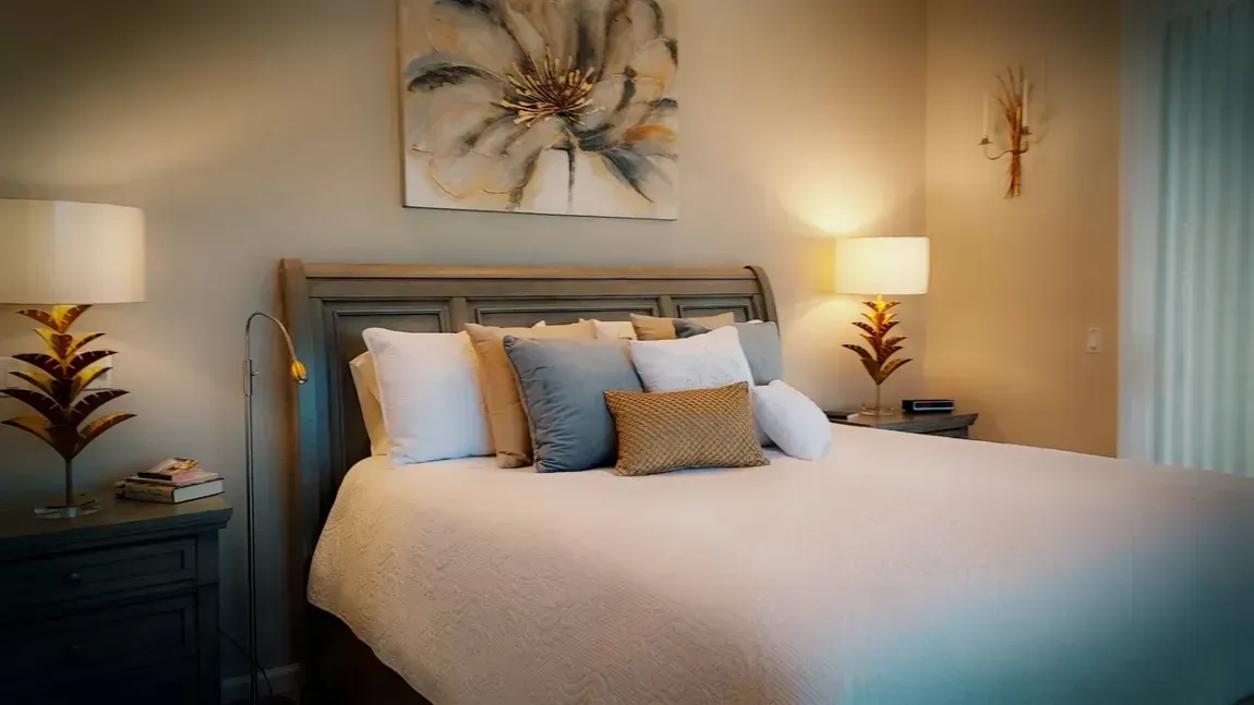 Bedroom with a bed, pillows, lamps, artwork, and nightstands. Soft, warm lighting and neutral color palette.