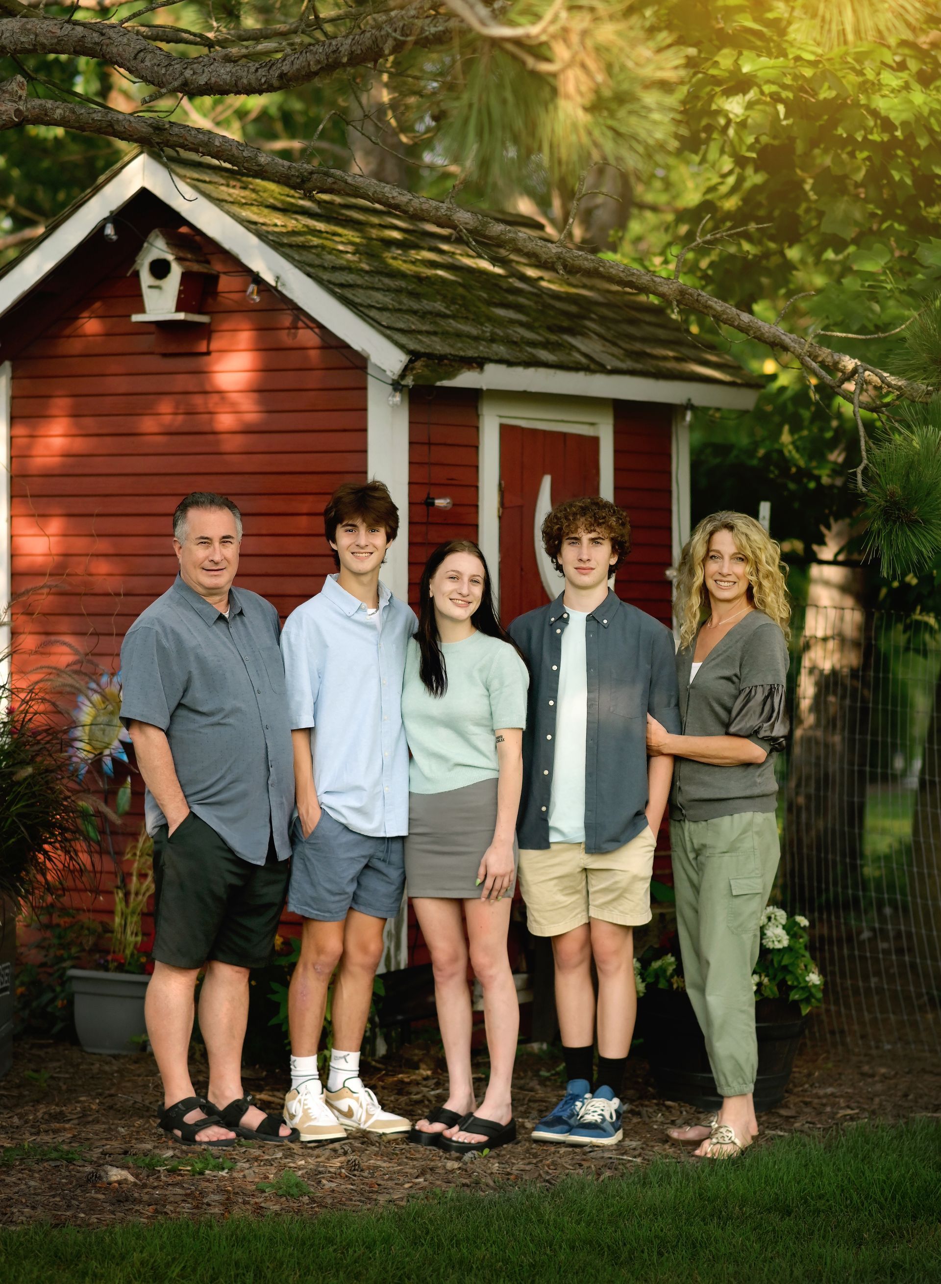 Five people standing together in front of a small red cabin in a wooded area