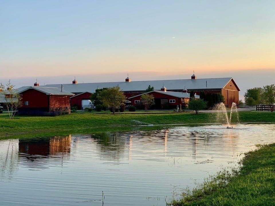 Red barns by a pond at sunset, with a fountain spraying water and reflections on the surface