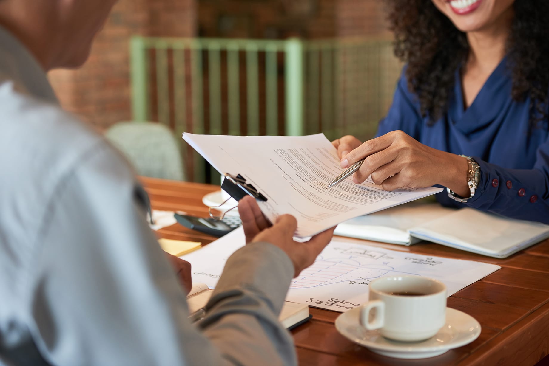 Two people discussing documents at a wooden table with a coffee cup in a bright office.