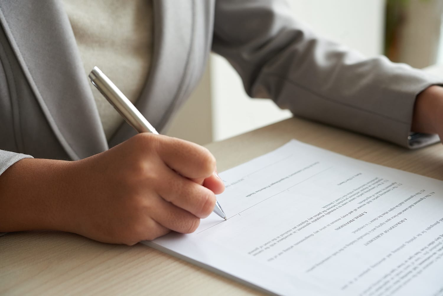 Hand writing on a document at a desk with a pen, wearing a light gray blazer