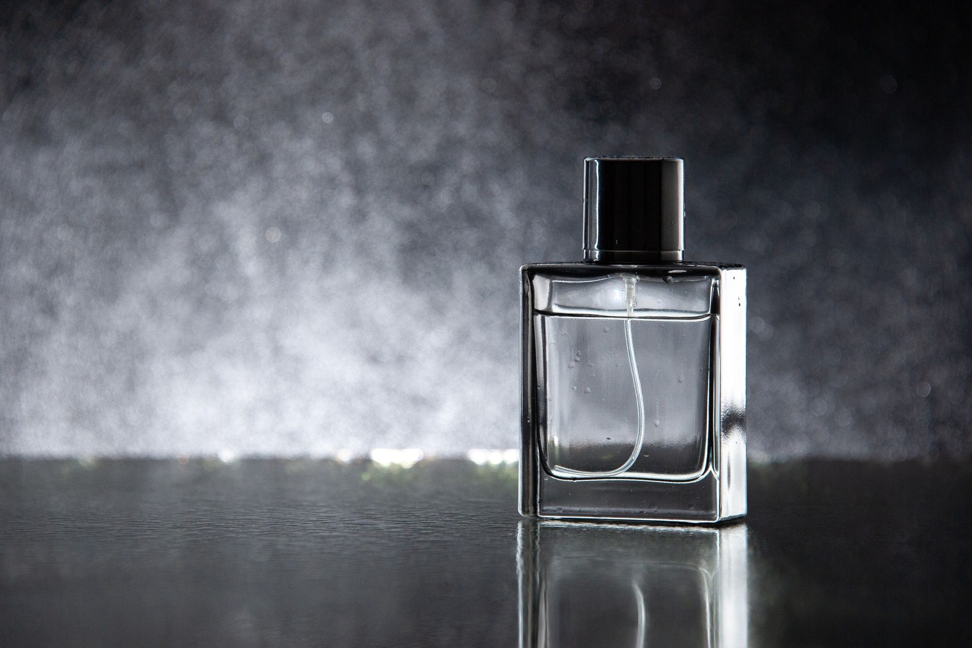 Frasco de perfume rectangular transparente con tapón negro, sobre superficie reflectante, rodeado de niebla.