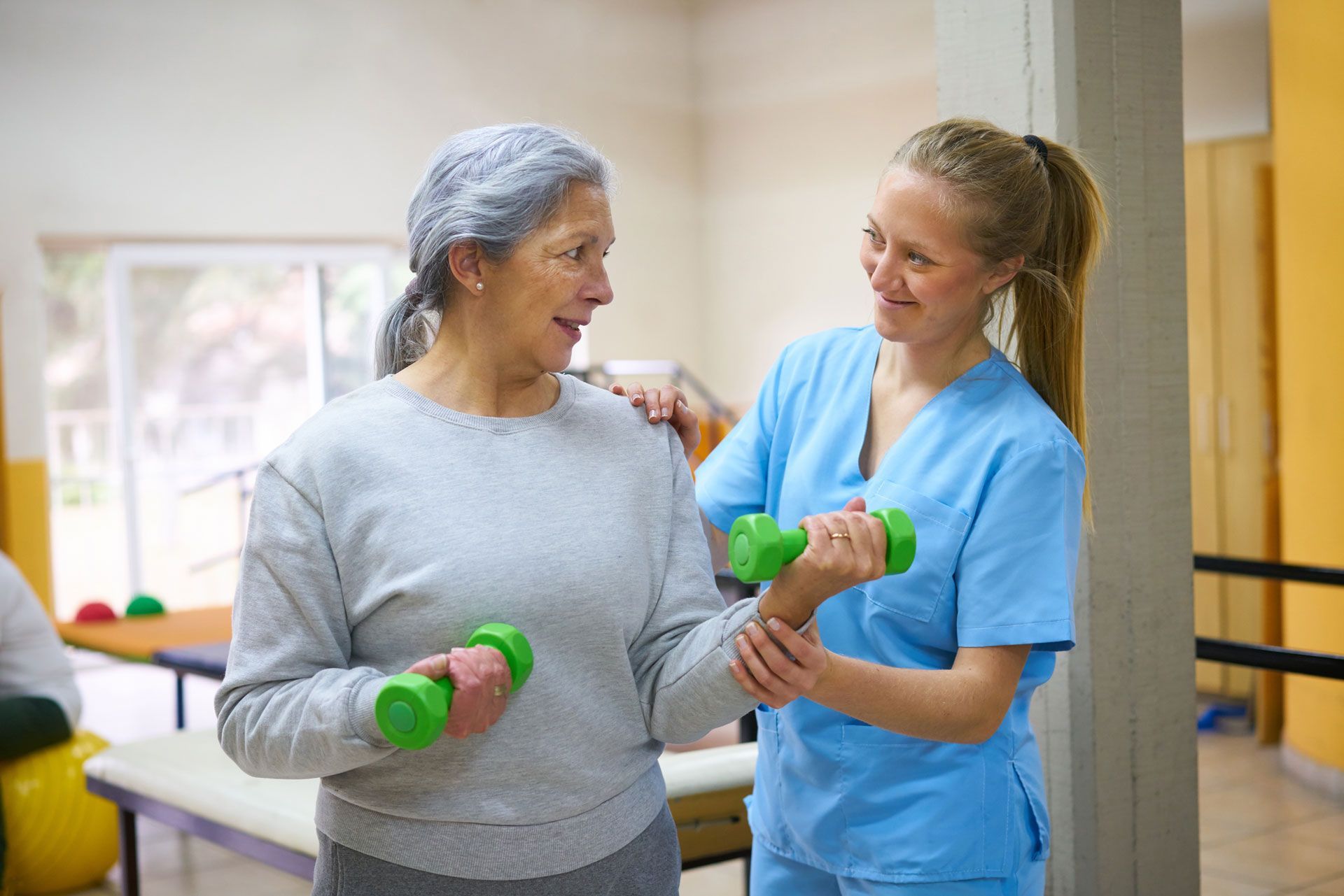 Therapist assisting a patient with arm exercises, holding green weights, in a light-filled therapy room.