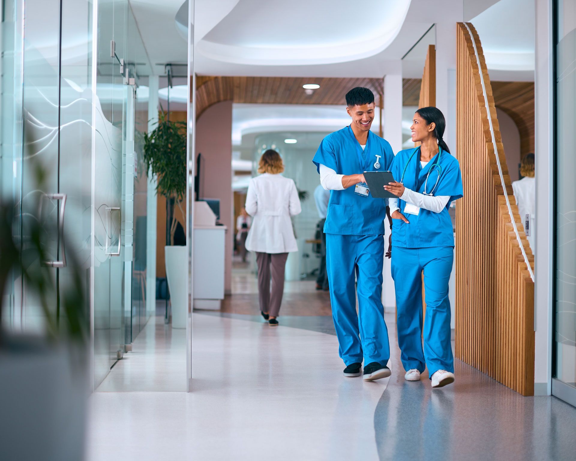 Two medical professionals in blue scrubs walking and discussing a chart in a brightly lit hospital hallway.