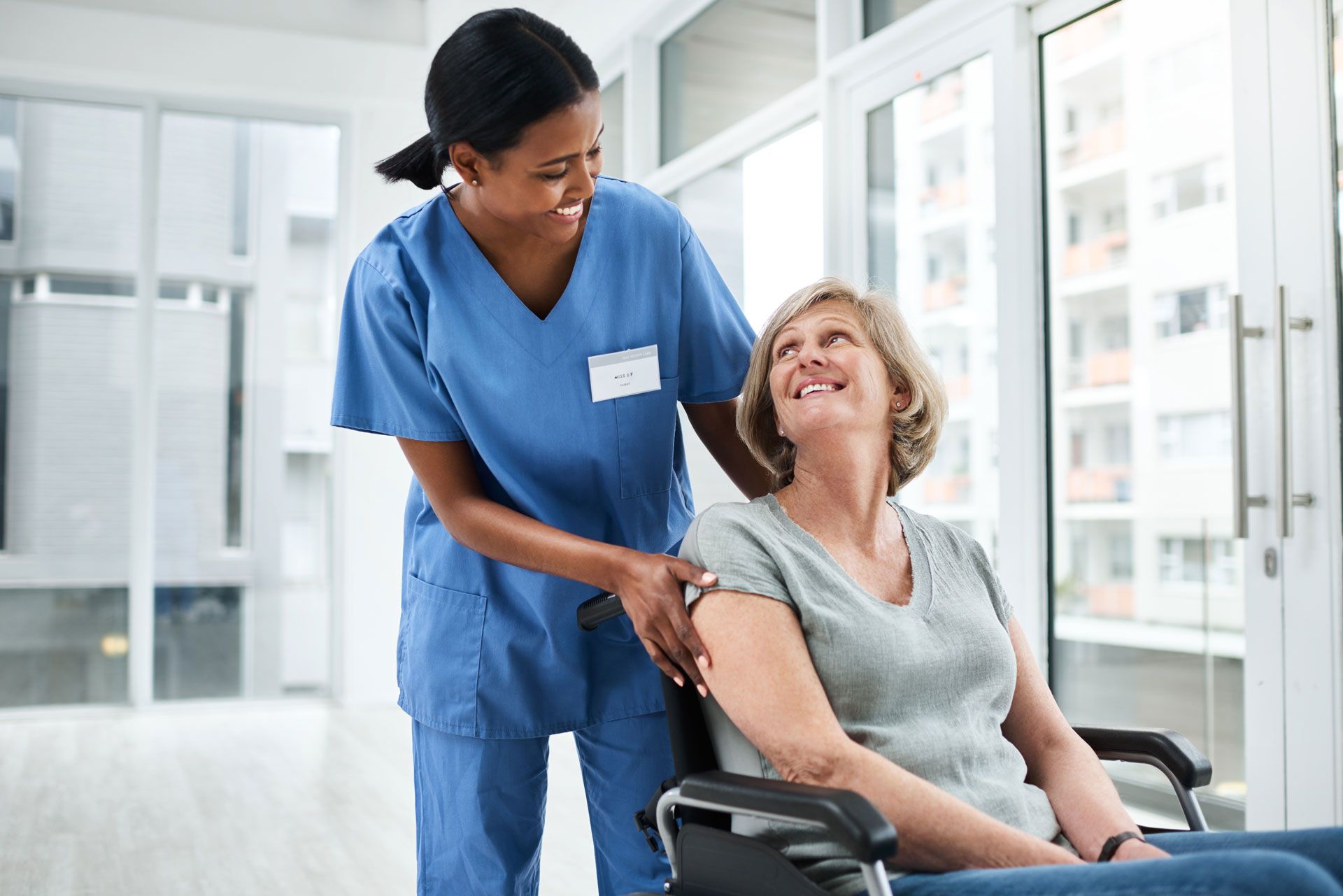Nurse in blue scrubs smiling while pushing a woman in a wheelchair through a bright hallway.