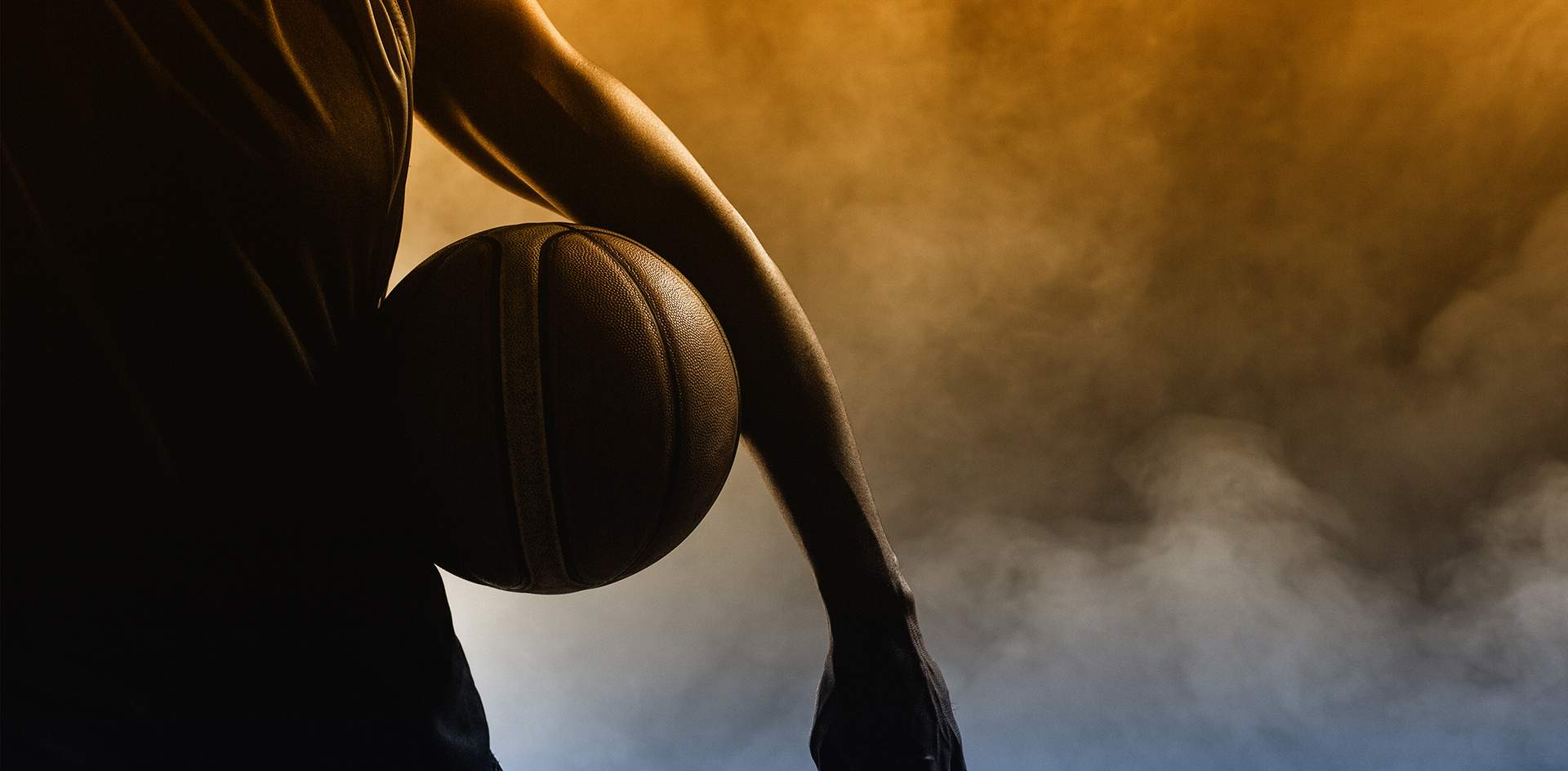 A person holding a basketball, silhouetted against a smoky, orange and yellow background.
