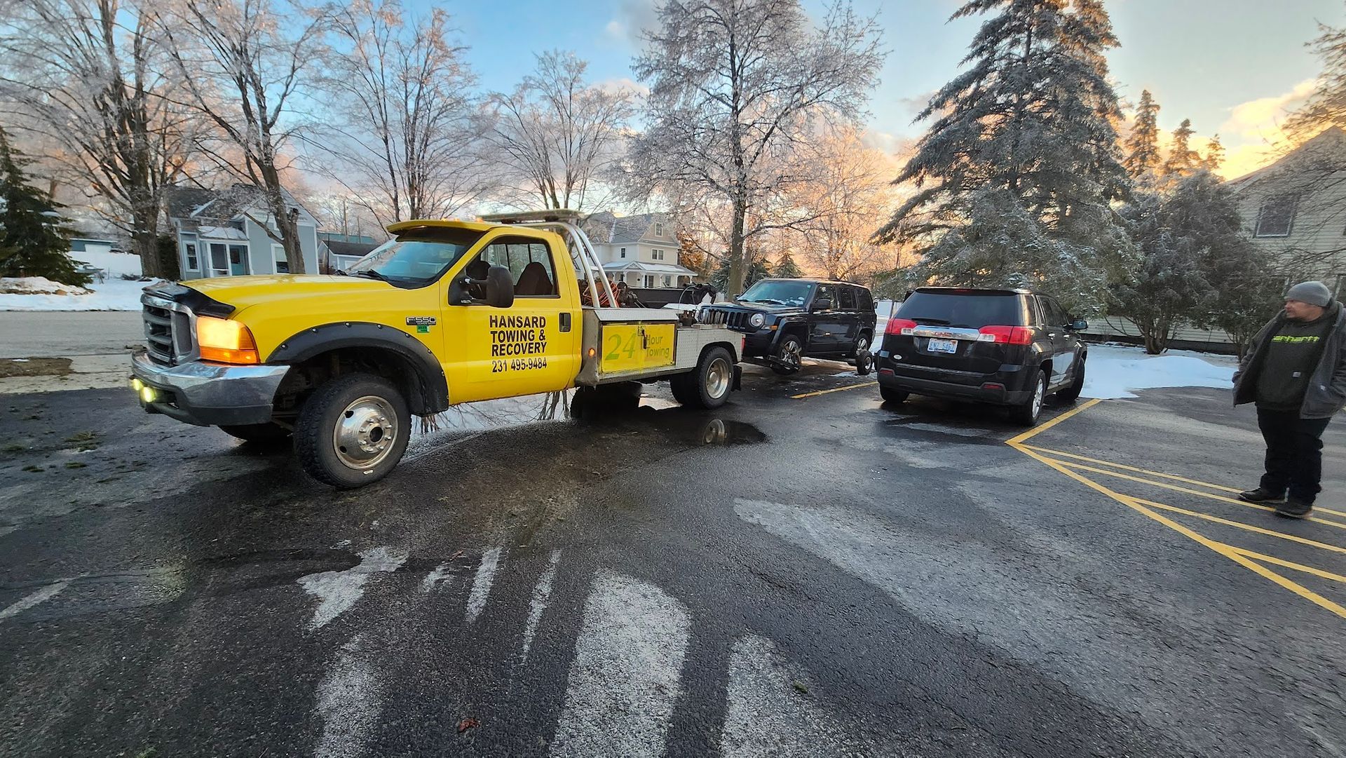A yellow tow truck is hooking up a black SUV in a parking lot. A person in a dark coat watches in a wintery, sunny setting.