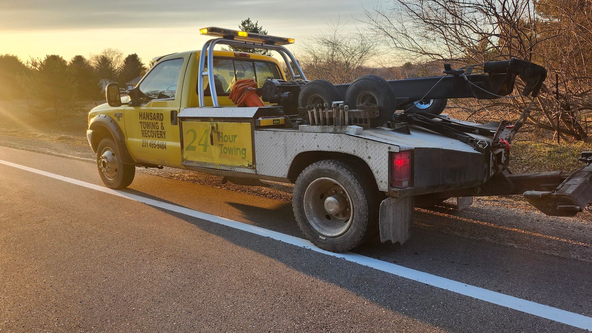 Yellow tow truck parked on the side of a road, facing away from the viewer. The sky is lit by the sun.