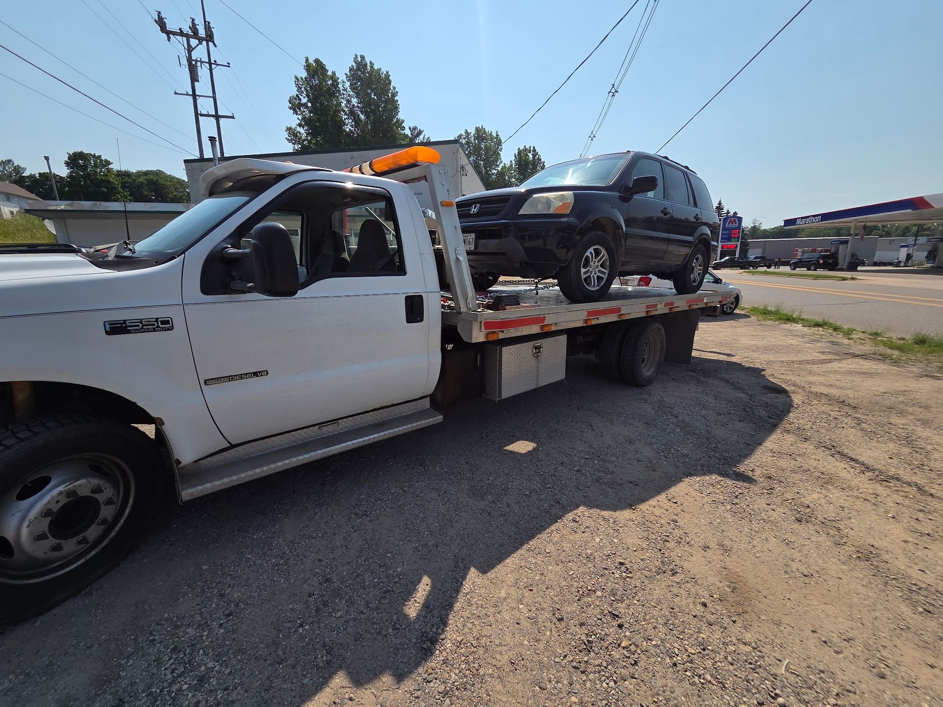 A black SUV is being towed on a flatbed tow truck. The truck is white and parked on a gravel surface on a sunny day.