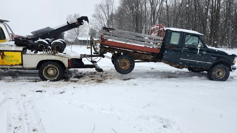A tow truck pulling a dark blue pickup truck with a wooden flatbed out of snowy terrain.