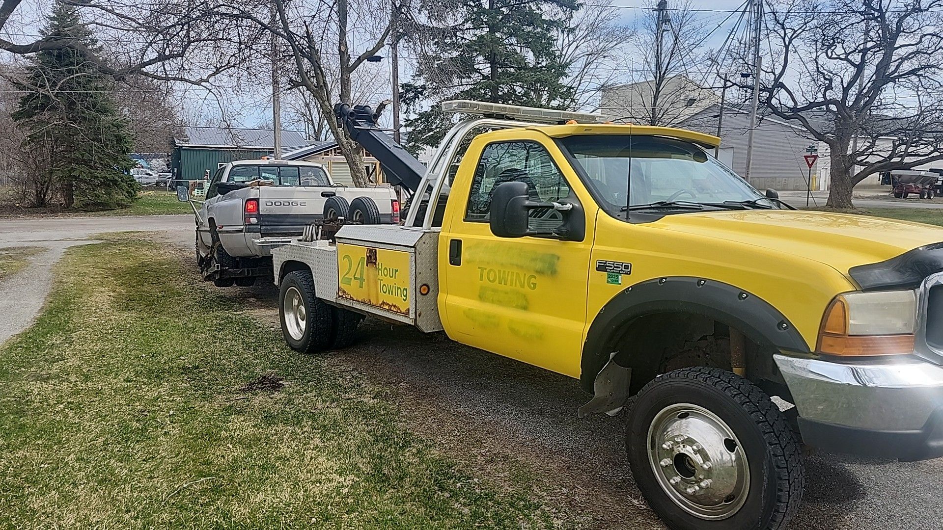 Yellow tow truck towing a silver pickup truck on a grassy area. The tow truck is parked next to a tree-lined street.