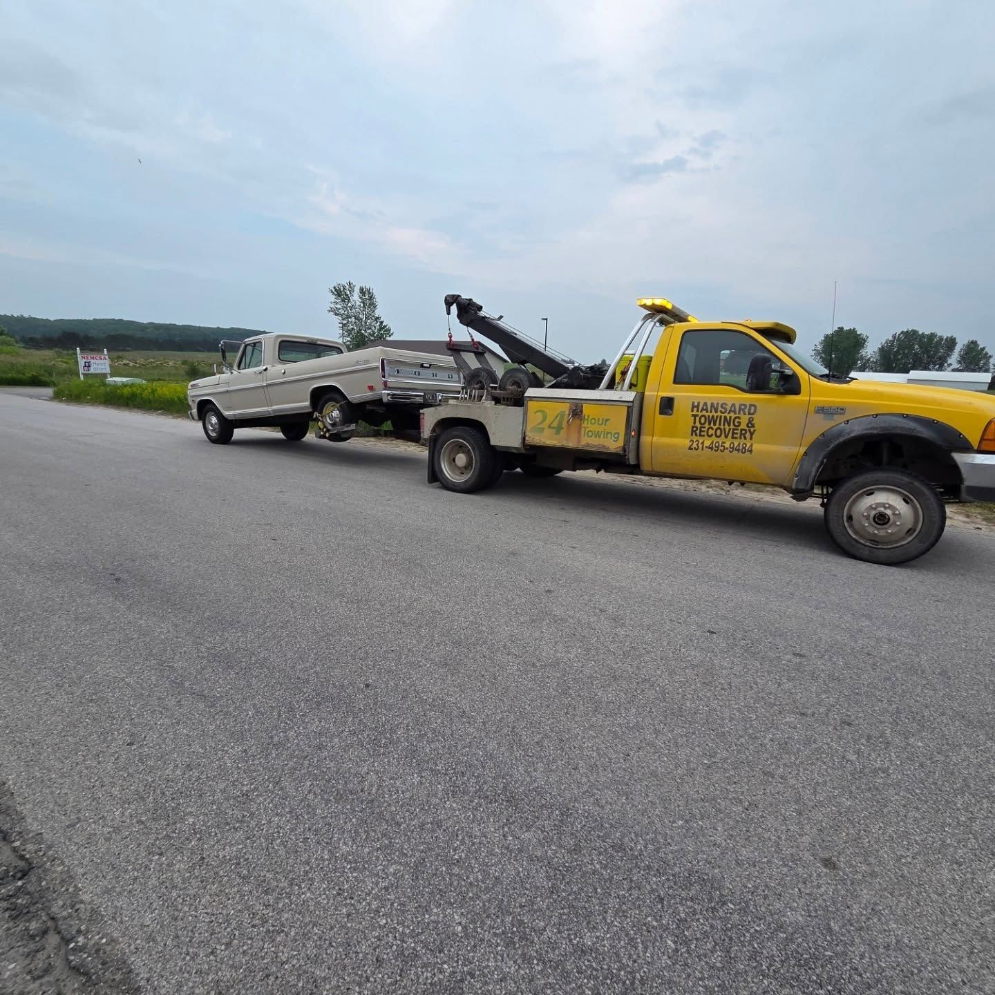 A yellow tow truck towing a white pickup truck on a gray road. The setting is a rural area.