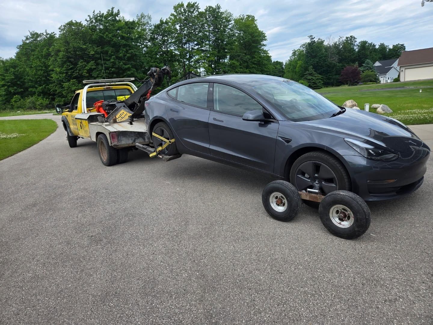 A gray Tesla Model 3 being towed by a yellow tow truck on a paved driveway. The car's front wheels are on a dolly.