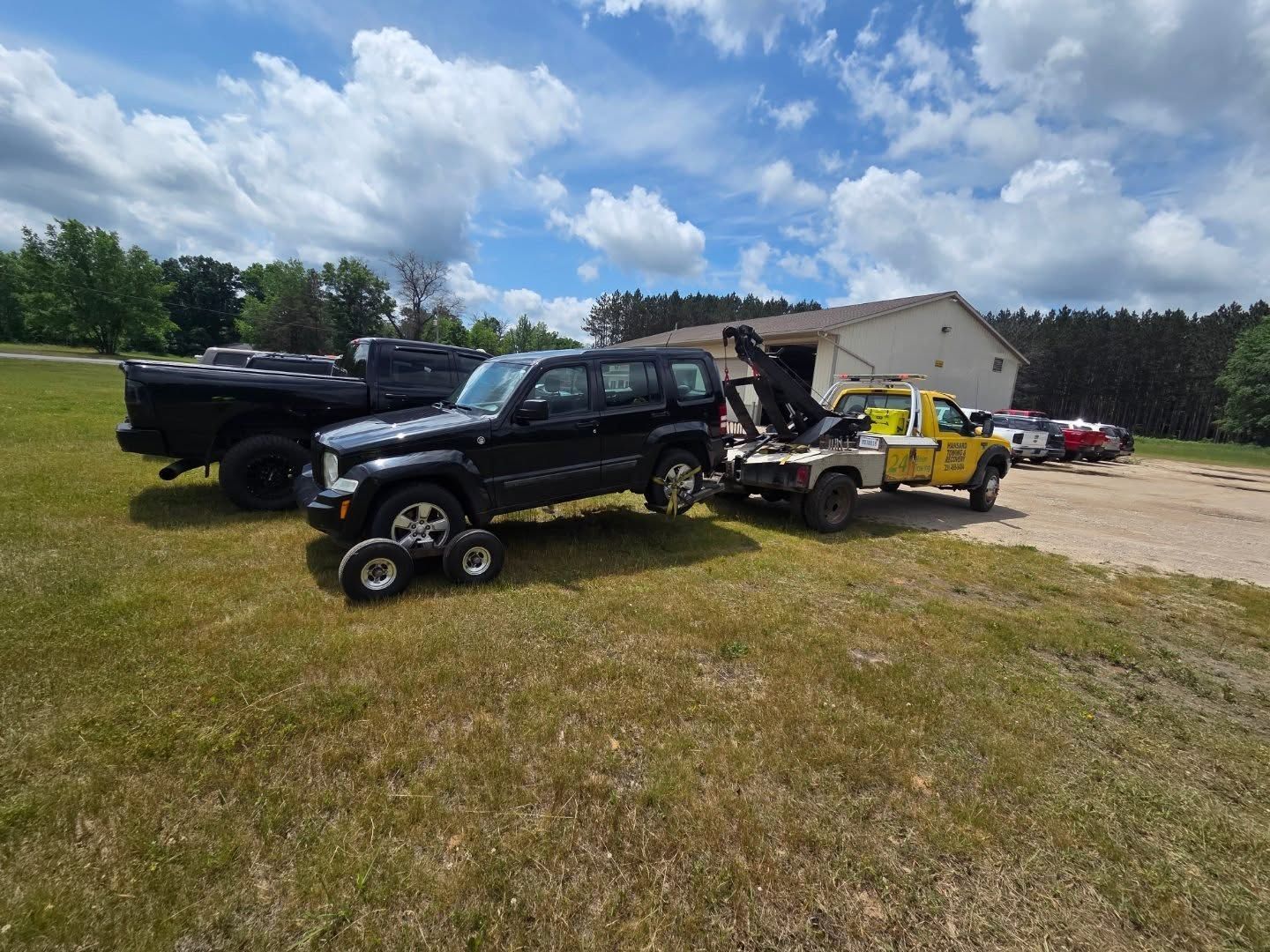 A black Jeep being towed by a yellow tow truck on a grassy field with other 