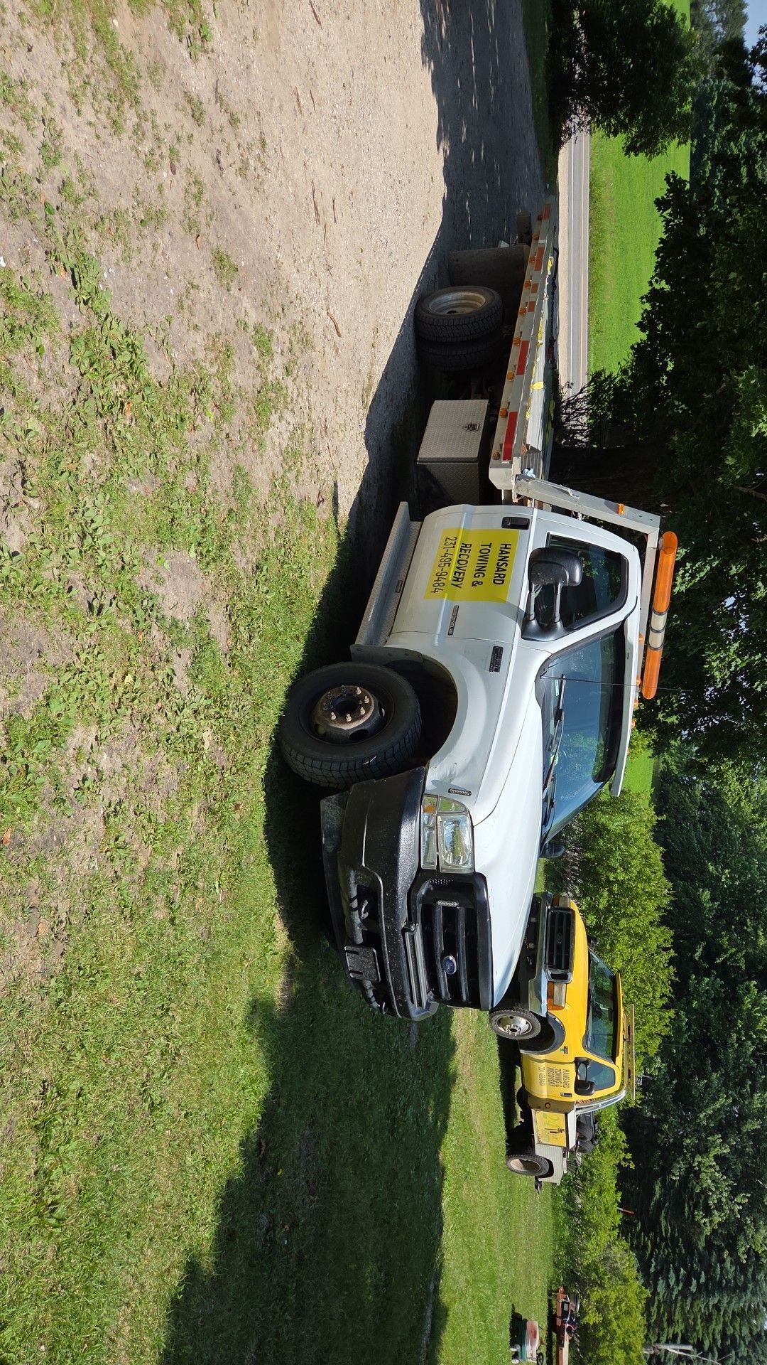 White flatbed tow truck parked on grass near a paved area.  Yellow truck visible in the background.