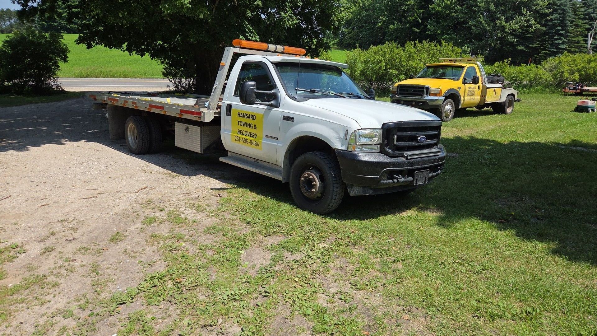 White tow truck parked on gravel and grass with a yellow tow truck in the background, in front of trees.