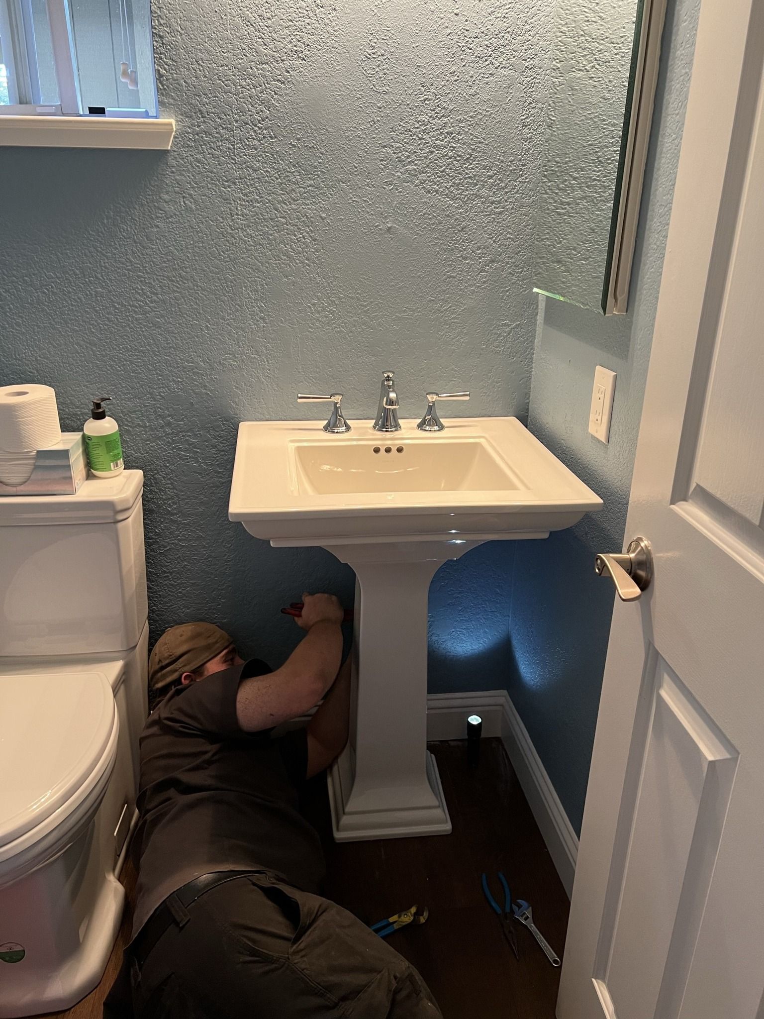 Person working on plumbing under a white pedestal sink in a bathroom with blue textured walls and toilet.