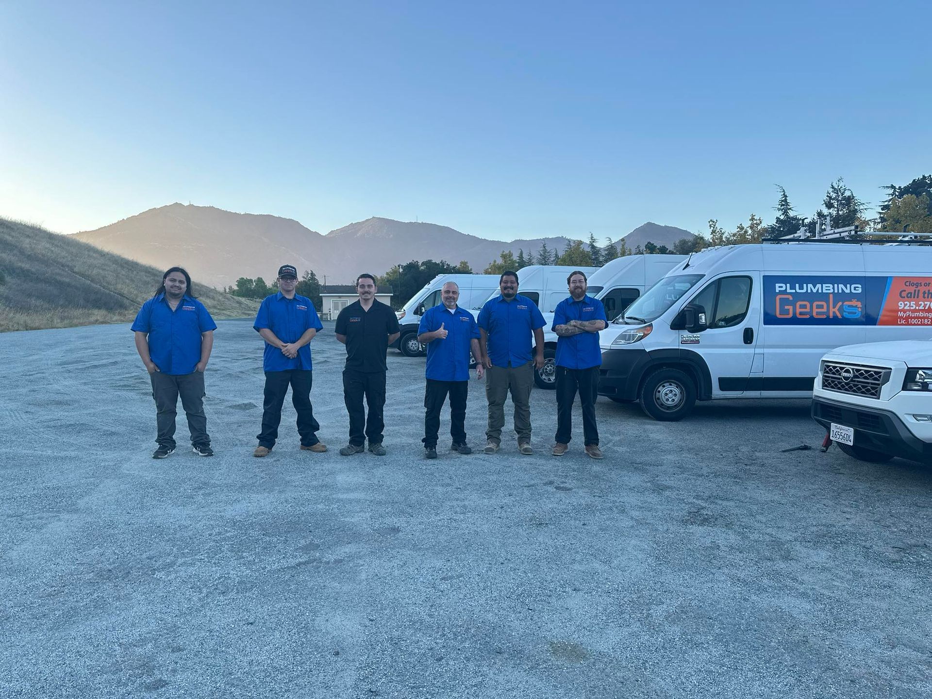 A group of service technicians in blue shirts pose with white vans in front of a mountain landscape.