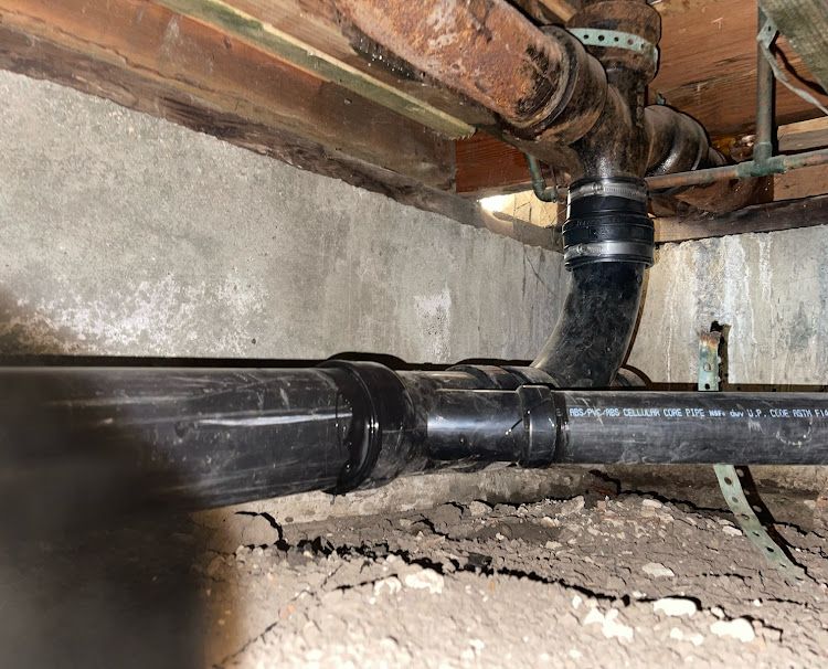 Black plumbing pipes and fittings under a building, against a concrete and wood backdrop.