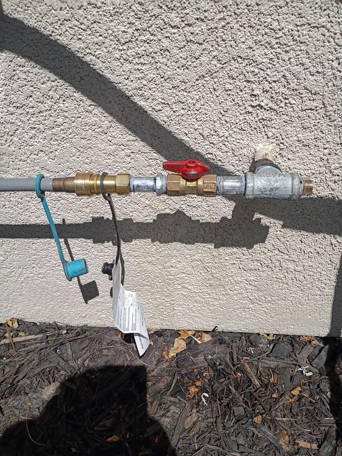 Gas line with red valve, attached to a gray pipe, on a stucco wall. Blue tag and tie.