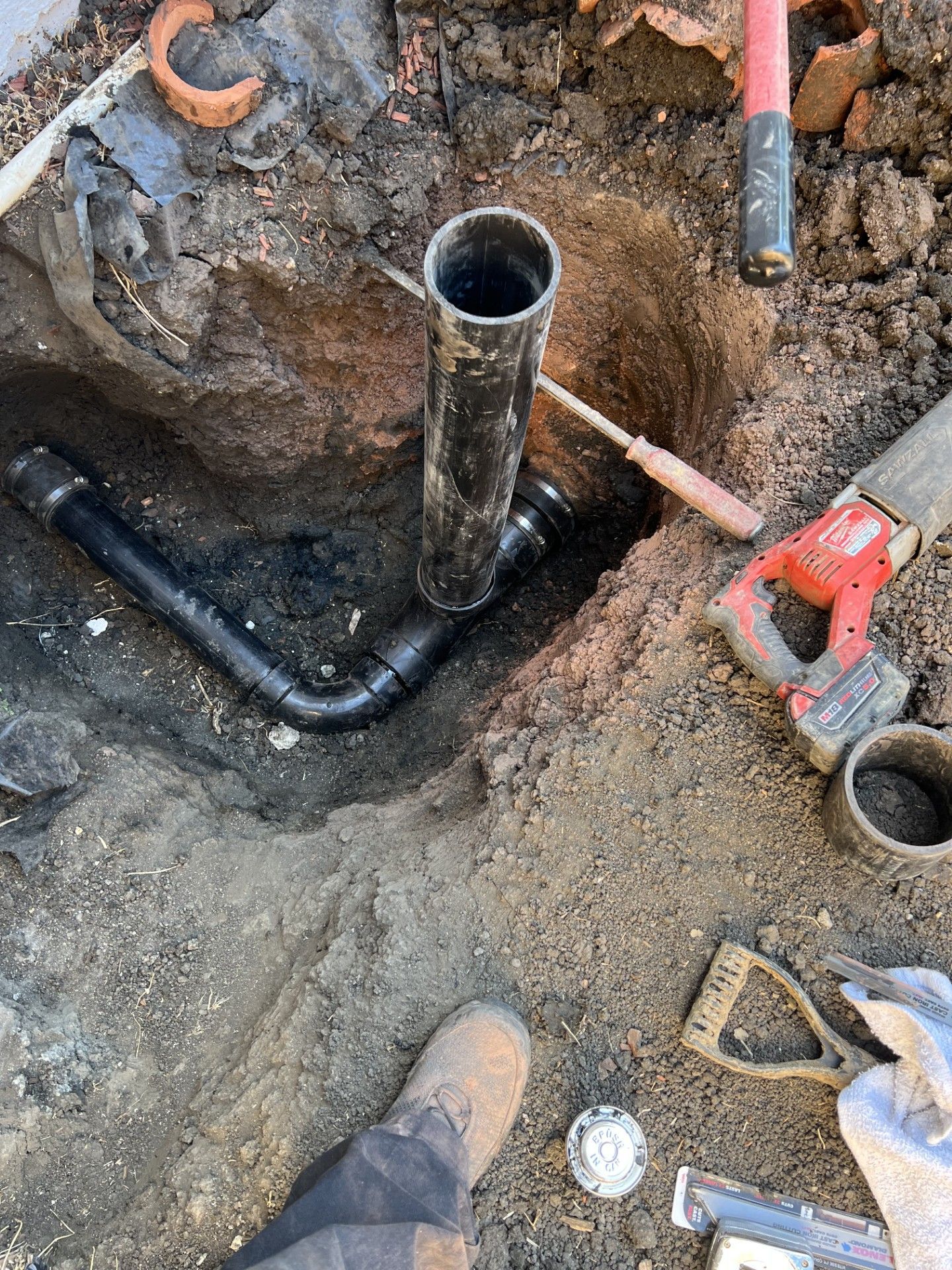 Black plumbing pipes in a dirt hole, with tools nearby; a person's muddy boot visible.