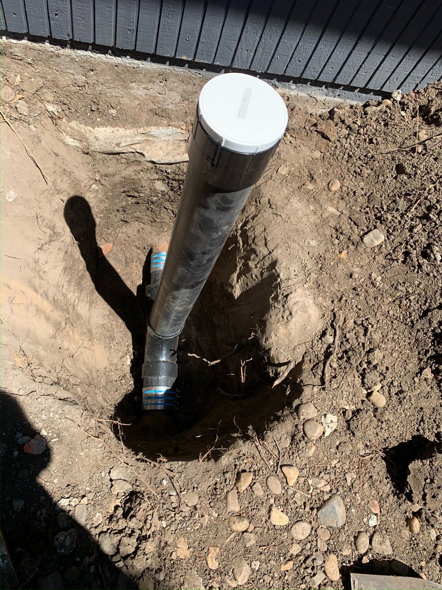 Black pipe with a white cap in a hole in the ground next to a dark fence.