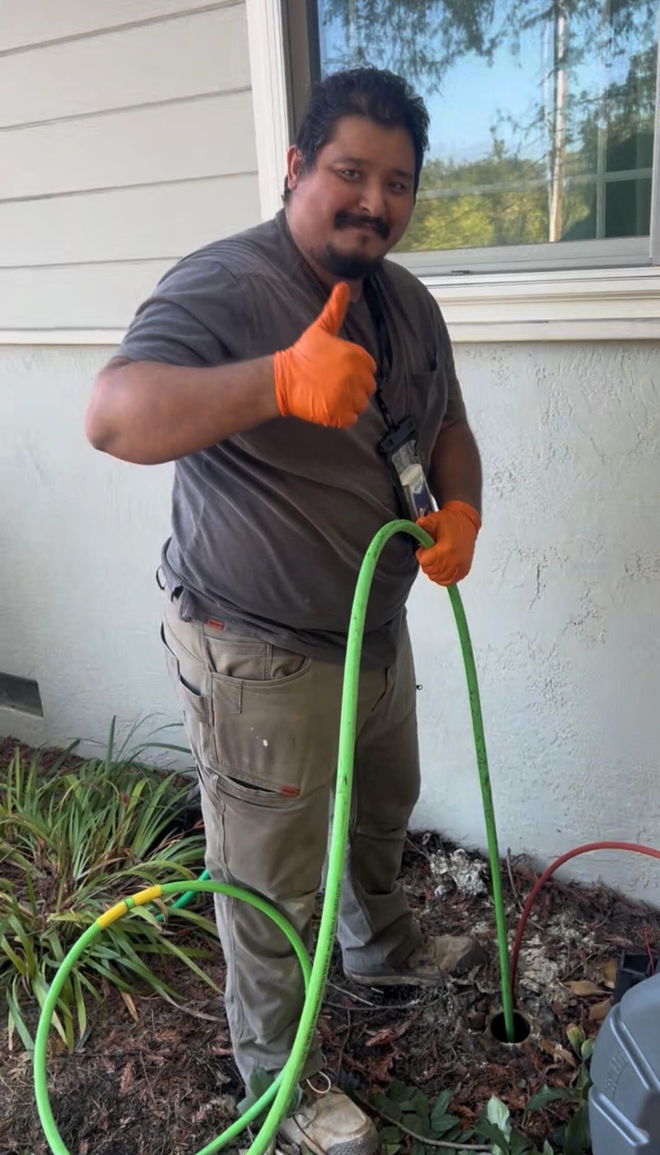 Man in work clothes giving thumbs up, holding hoses near ground, orange gloves.