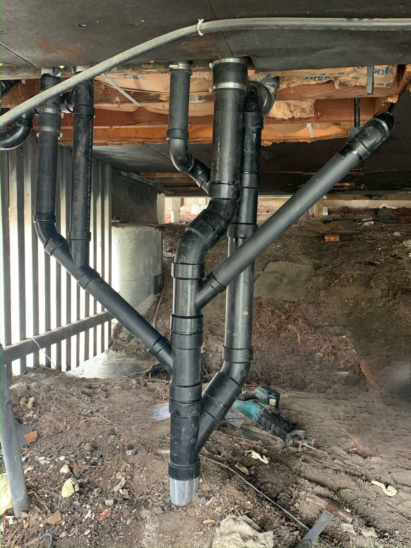 Black plumbing pipes converge and extend beneath a wooden structure, set in a dirt crawl space.